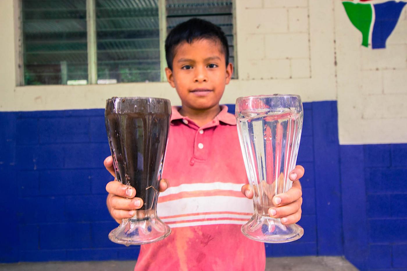 A local boy holds two glasses of water, demonstrating the difference between locally sourced water and a glass of water passed through the purifying filter brought to his community by The WaterVan project.