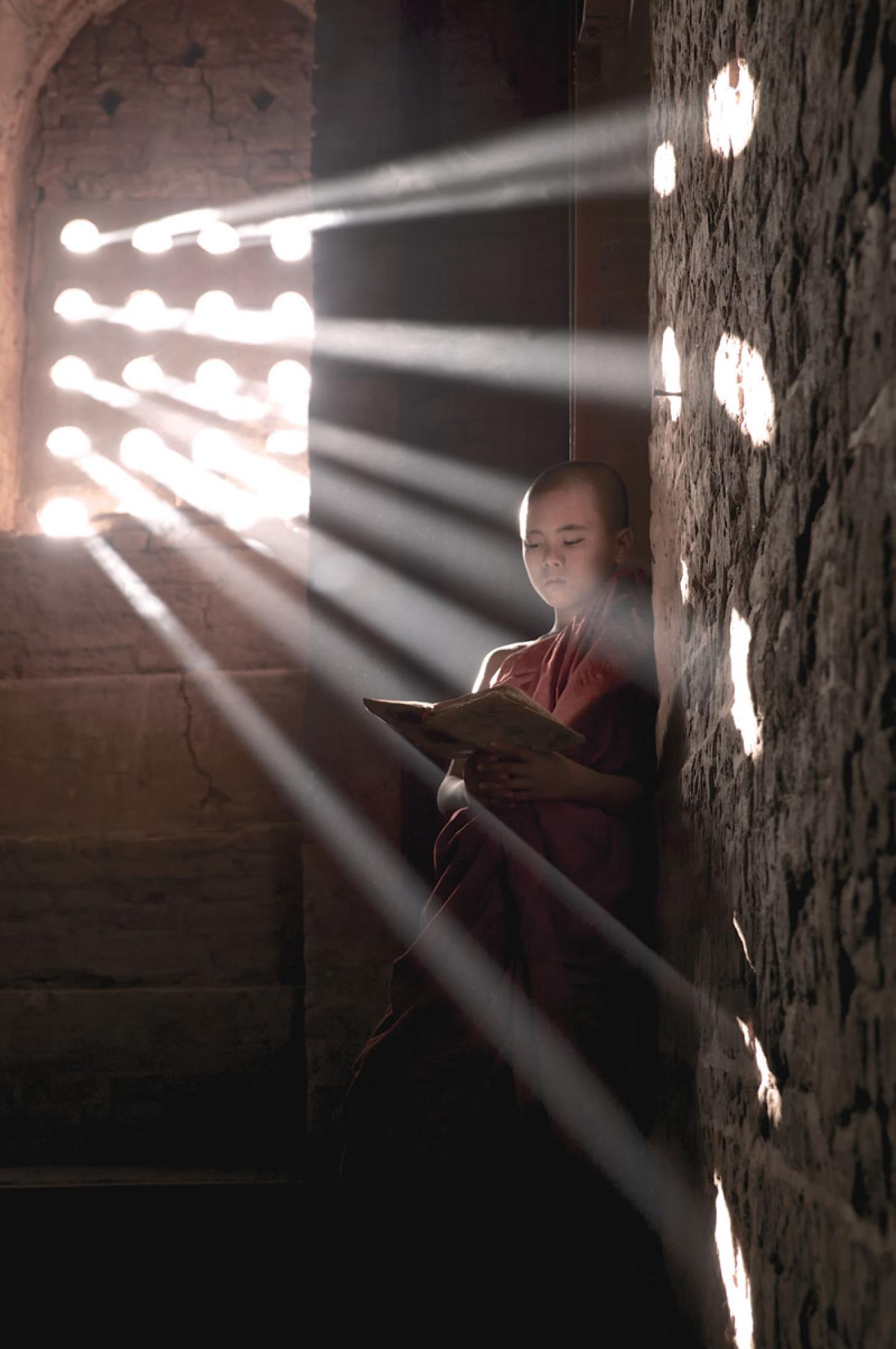 Bagan, Myanmar. Rays of light pierce through a nearby window falling on a novice Buddhist Monk and his book.