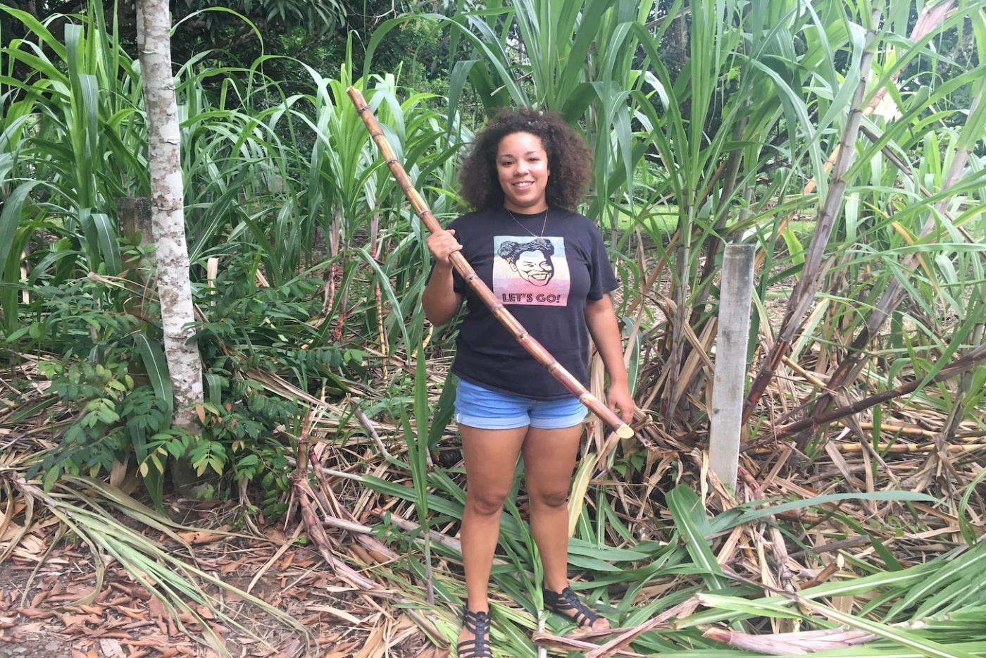 Marisa standing in a crop of sugar cane, holding a fresh cut piece.